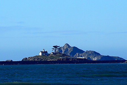 [The two-story house with the lighthouse on the third level sits on an island with one tree higher than the house and several rock hills higher than the house. The sky is clear with only some wisps of clouds in the distance.]